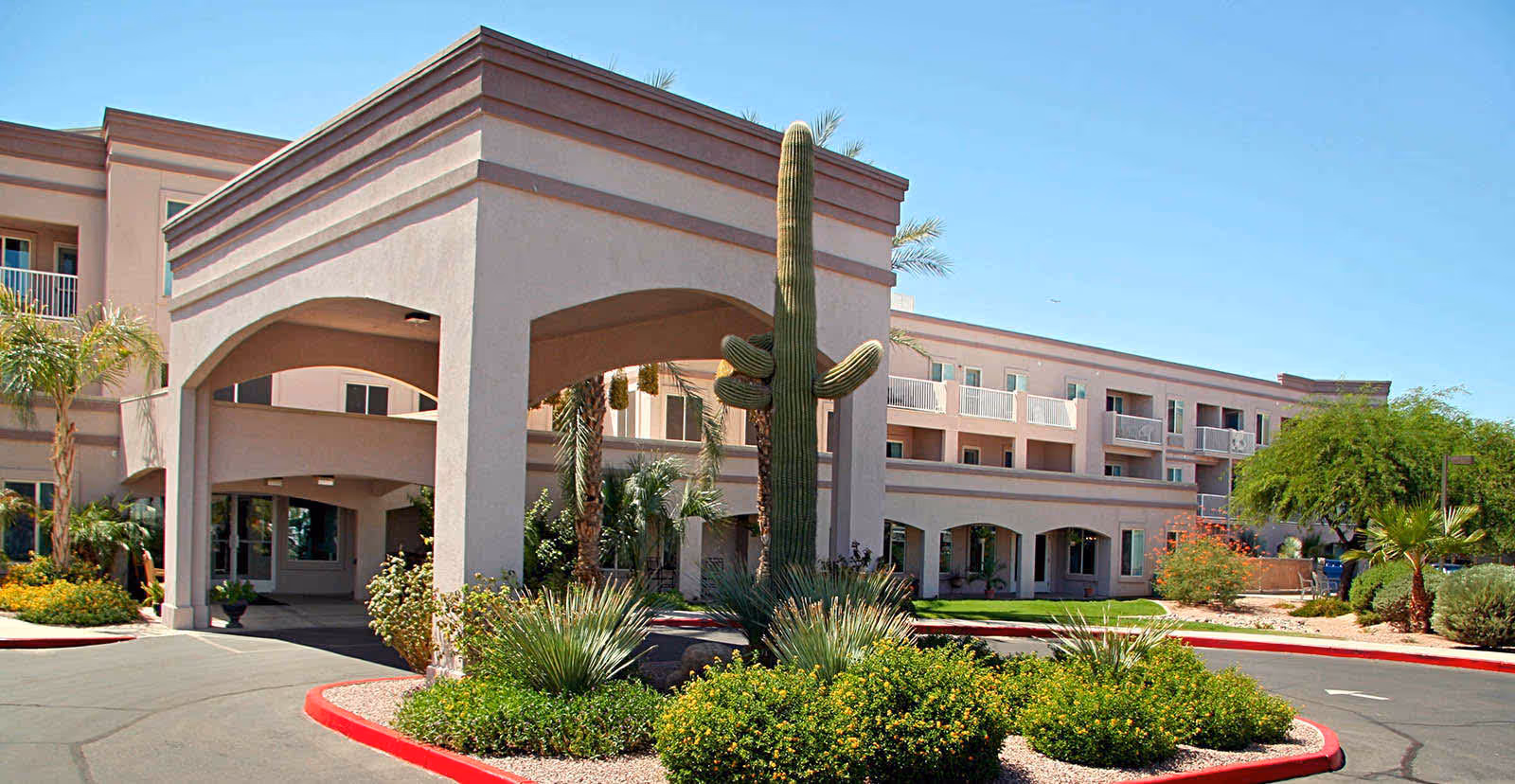 Exterior view of Holiday Vista de la Montana senior living facility showing a three-story building with balconies, a covered entrance with arches, desert landscaping including a large cactus, palm trees, and shrubs under a clear blue sky.