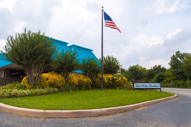 Exterior view of East Ridge Residence with turquoise roof, landscaped shrubs, flagpole, and sign.