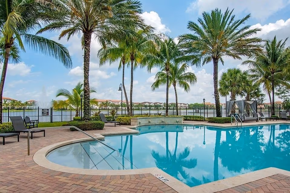 Outdoor swimming pool area with lounge chairs and palm trees surrounding it, a lake with a fountain in the background, and a partly cloudy sky.