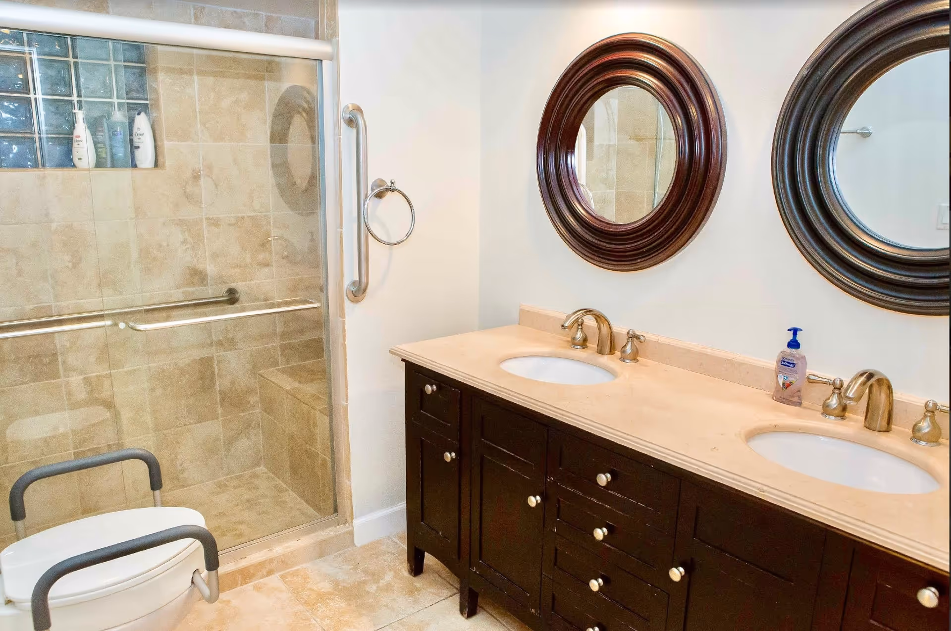 A bathroom with a double sink vanity featuring dark wood cabinets and beige countertop. Above the sinks are two round mirrors with dark wooden frames. To the left is a walk-in shower with beige tiles, a built-in bench, glass block window, and a glass door with a metal handle. In front of the shower is a white toilet with gray armrests.