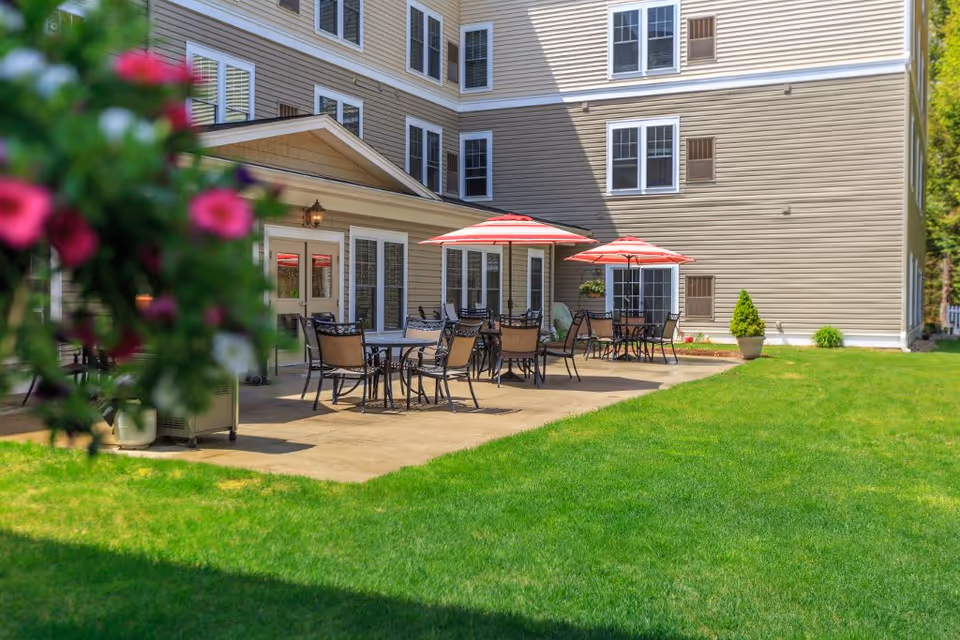Outdoor patio area at a senior living facility with several tables and chairs under red and white striped umbrellas, adjacent to a beige multi-story building with multiple windows. There is a well-maintained green lawn in the foreground and some blurred flowers on the left side of the image.
