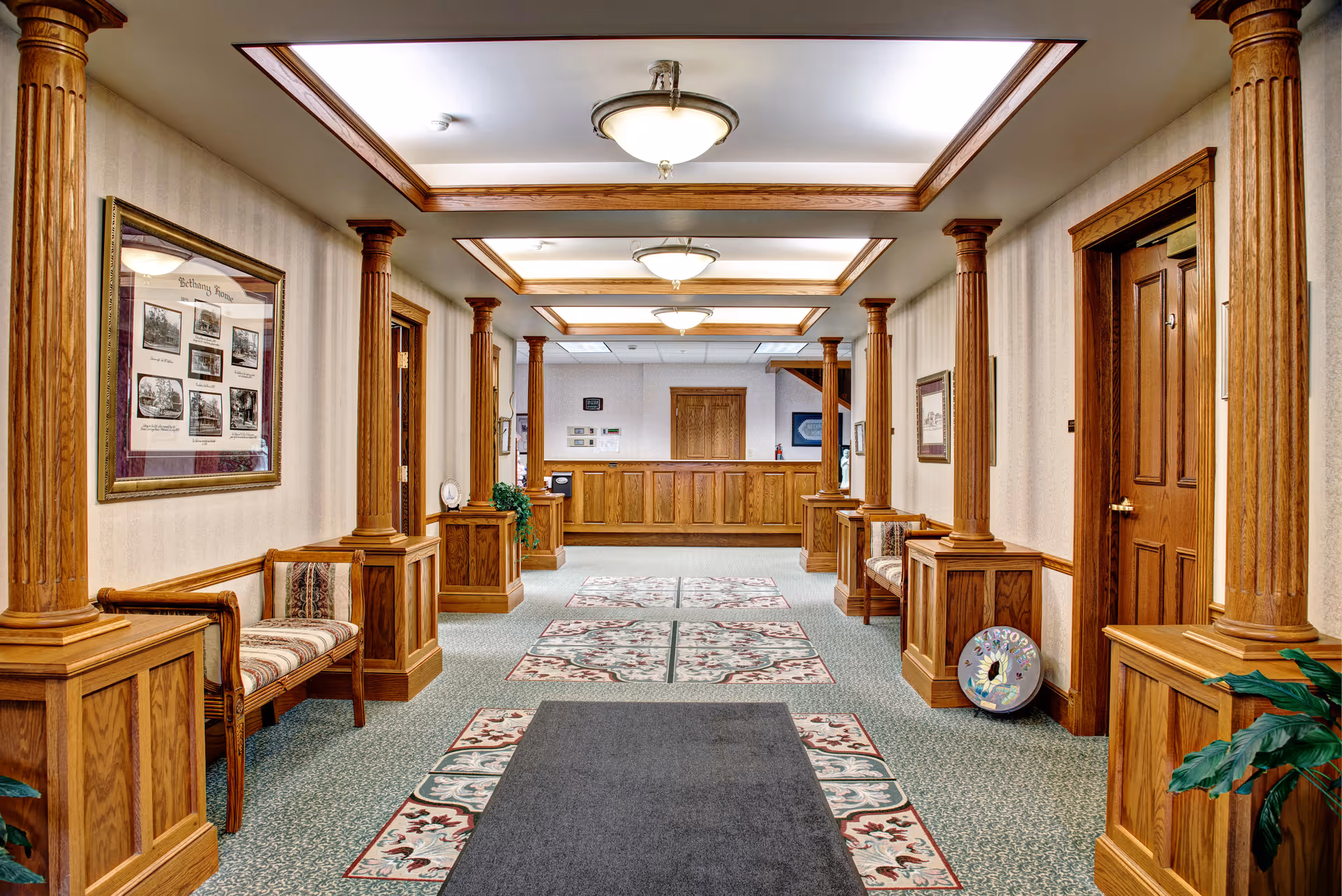 Interior hallway of Bethany Home Retirement Center featuring wooden columns and paneling, patterned carpet with floral designs, cushioned wooden benches along the walls, framed pictures, and ceiling lights with wooden trim. A reception desk is visible at the far end of the hallway.