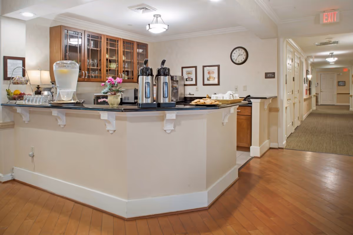 Interior common area with a serving counter featuring coffee urns, a water dispenser, pastries and glass-front cabinets, with a hallway beyond.