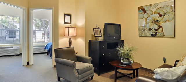A cozy living room area with a gray armchair, a round wooden coffee table with a plant and remote control, a black dresser with an old-style TV and framed pictures, and a large floral painting on the wall. In the background, there is a bedroom with a bed covered in blue bedding, visible through an open doorway.