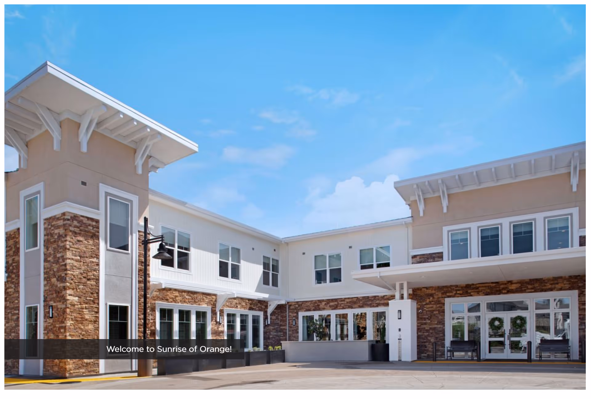 Exterior view of Sunrise of Orange senior living facility showing a modern two-story building with stone and beige siding, multiple windows, and a covered entrance under a clear blue sky.