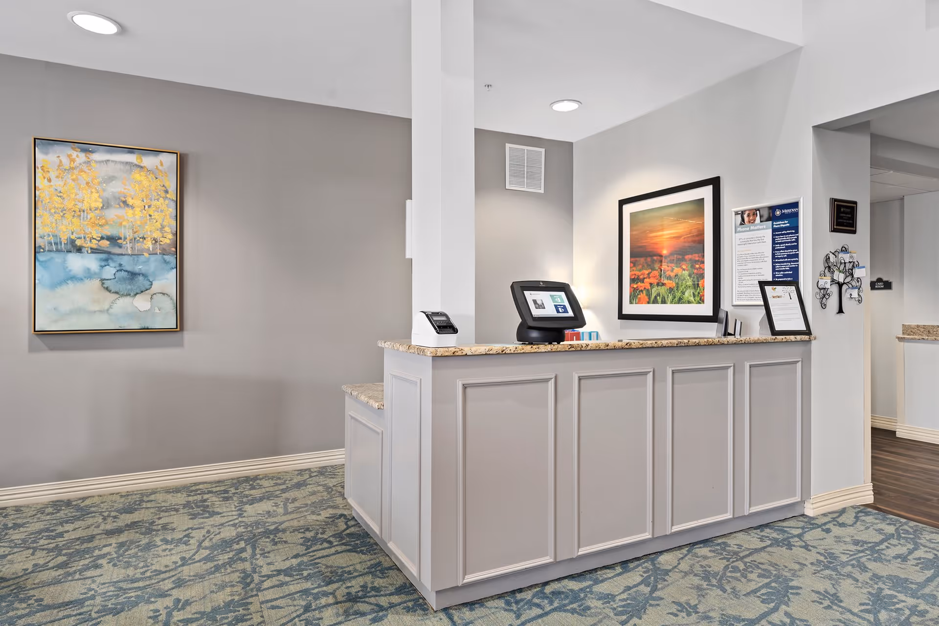 Reception desk area with a granite countertop in a senior living facility. Behind the desk is a framed picture of a sunset over a field of flowers, a digital check-in device, and informational posters on the wall. To the left, there is a wall with a framed painting of yellow trees over a blue and gray background. The floor is covered with a patterned carpet in shades of blue and gray.