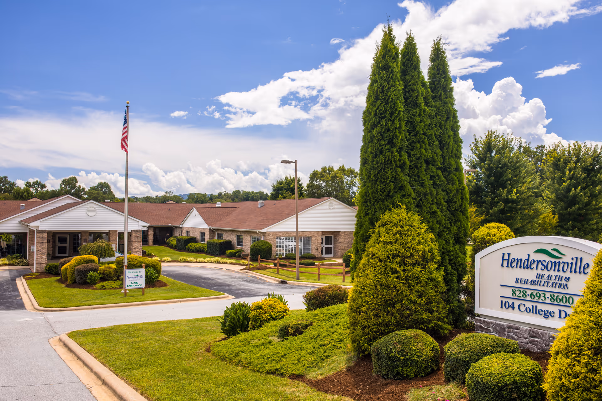 Exterior view of Hendersonville Health & Rehab facility with a driveway, manicured bushes, tall trees, and an American flag on a flagpole under a partly cloudy blue sky.
