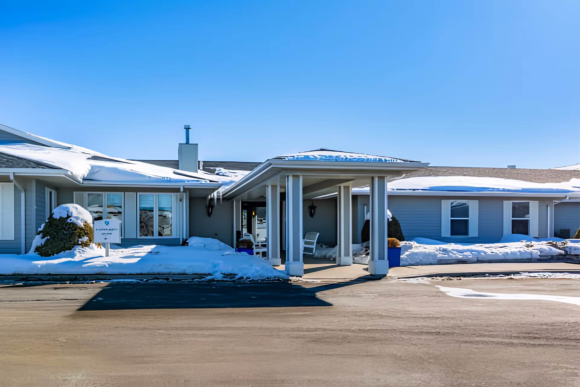 Exterior view of Elison Assisted Living of Minot building entrance with snow on the ground and roof under a clear blue sky. The entrance features a covered walkway supported by white columns and a few chairs are visible under the covered area.