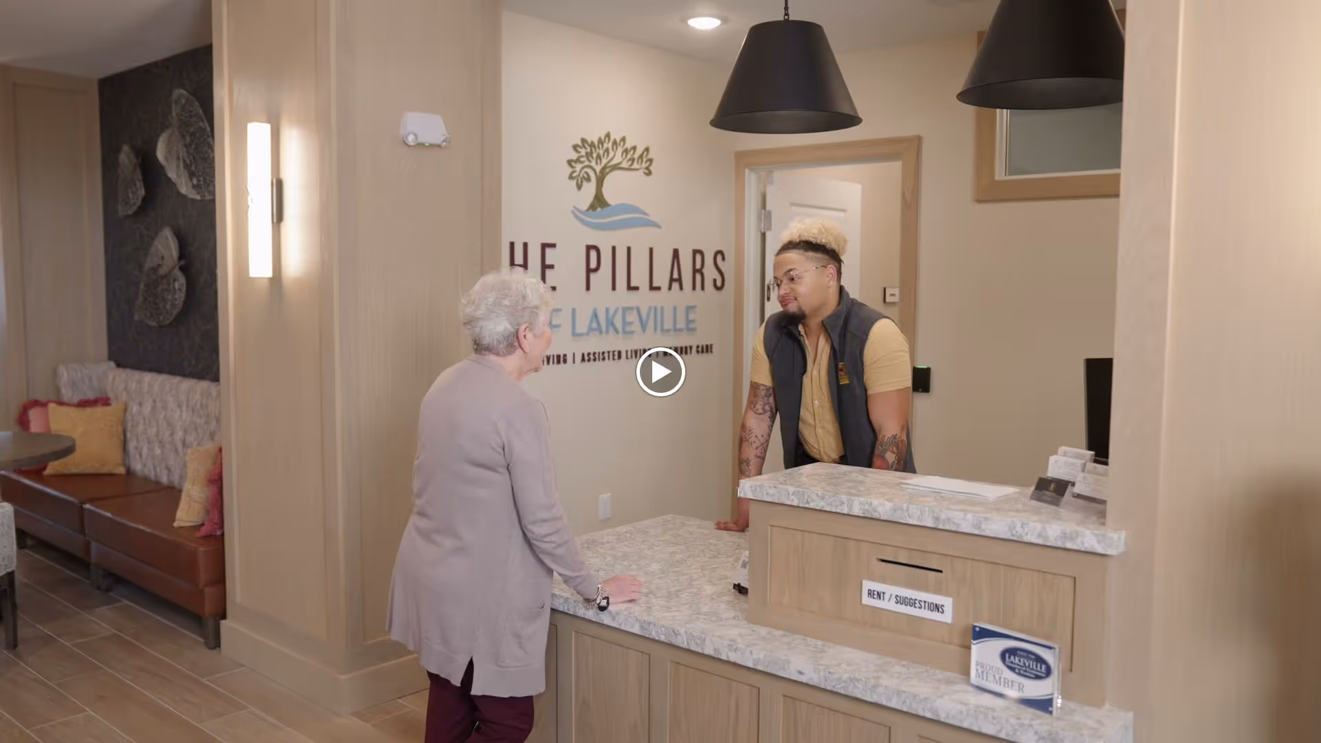 An elderly woman talking to a staff member at the reception desk inside The Pillars of Lakeville senior living facility. The reception area has a marble countertop, wooden paneling, and pendant lights. There is a seating area with cushions visible to the left.