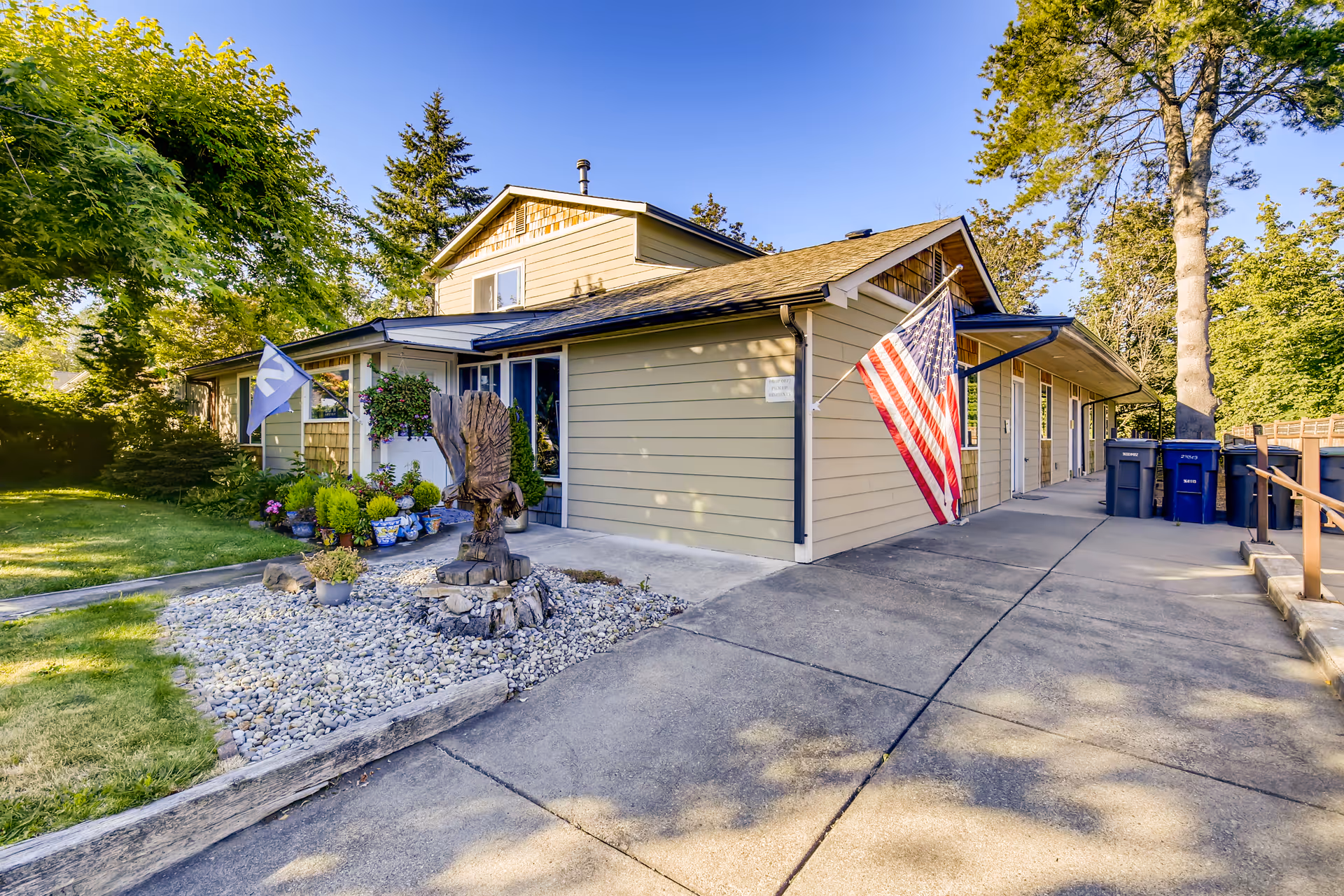 Beige single-story residential building with an American flag, driveway, and a small rock garden with a wooden sculpture.
