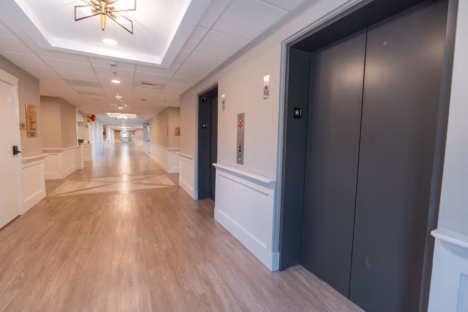 A long, well-lit hallway in a senior living facility with light wood flooring and beige walls. On the right side, there are two closed dark gray elevator doors marked with a star and the number 1. The ceiling has recessed lighting and a decorative light fixture. There are signs and balloons visible further down the hallway.
