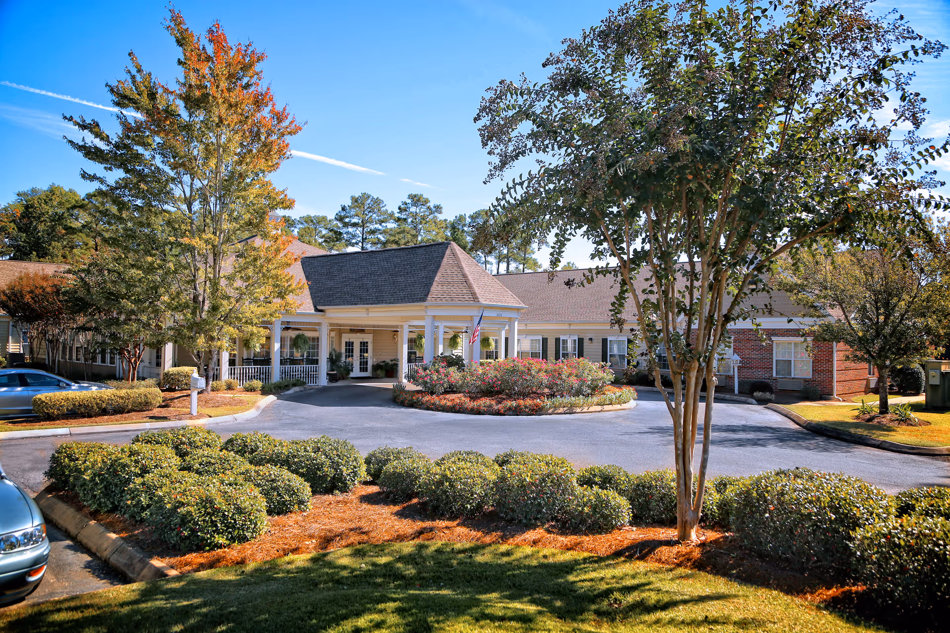 Front exterior view of a senior care facility named Woodleaf Senior Care, featuring a circular driveway with landscaped bushes and trees, a covered entrance with white pillars, and an American flag near the entrance. The building has a combination of beige siding and red brick with multiple windows.