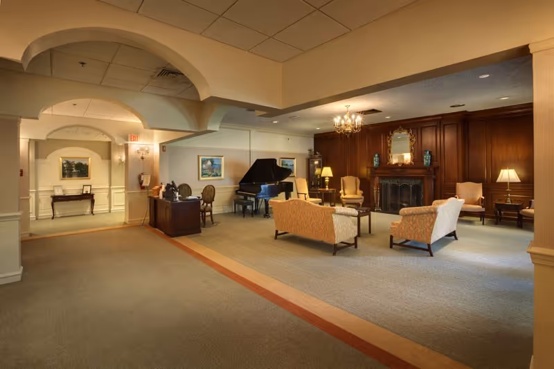 Spacious senior living common room with upholstered seating arranged around a fireplace, a grand piano, chandelier, and arched ceiling details.