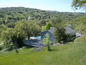 A scenic view of a countryside area with green grassy hills and dense trees surrounding a small building with a fenced yard and a swimming pool.