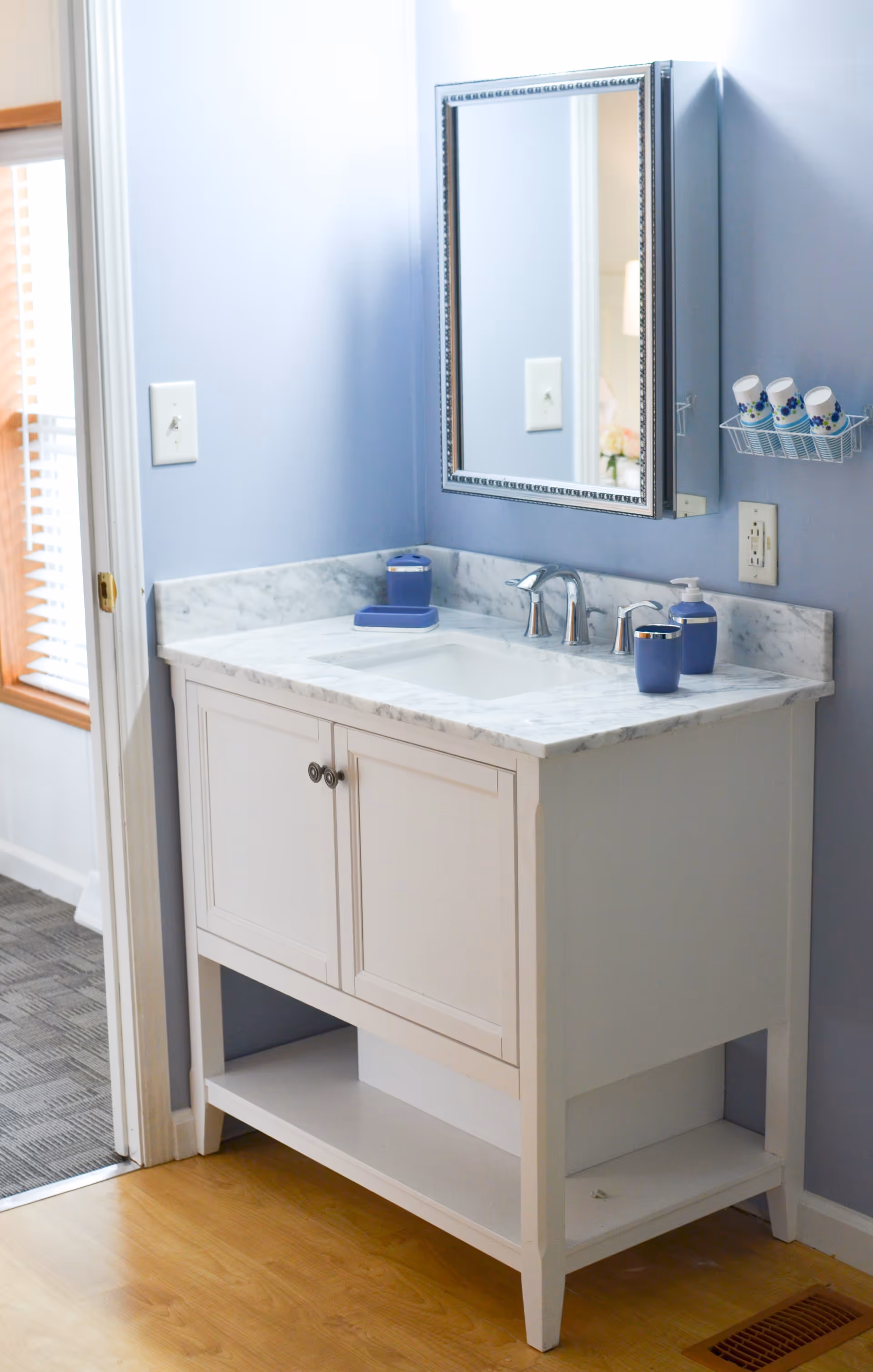A bathroom vanity with a white cabinet and marble countertop, featuring a sink with a silver faucet. Above the vanity is a rectangular mirror with a decorative frame. On the countertop are blue soap dispensers and a toothbrush holder. The walls are painted light blue, and there is a small wire rack mounted on the wall holding three cups. The floor is wooden, and a vent is visible near the vanity. A doorway with a carpeted floor is seen to the left.
