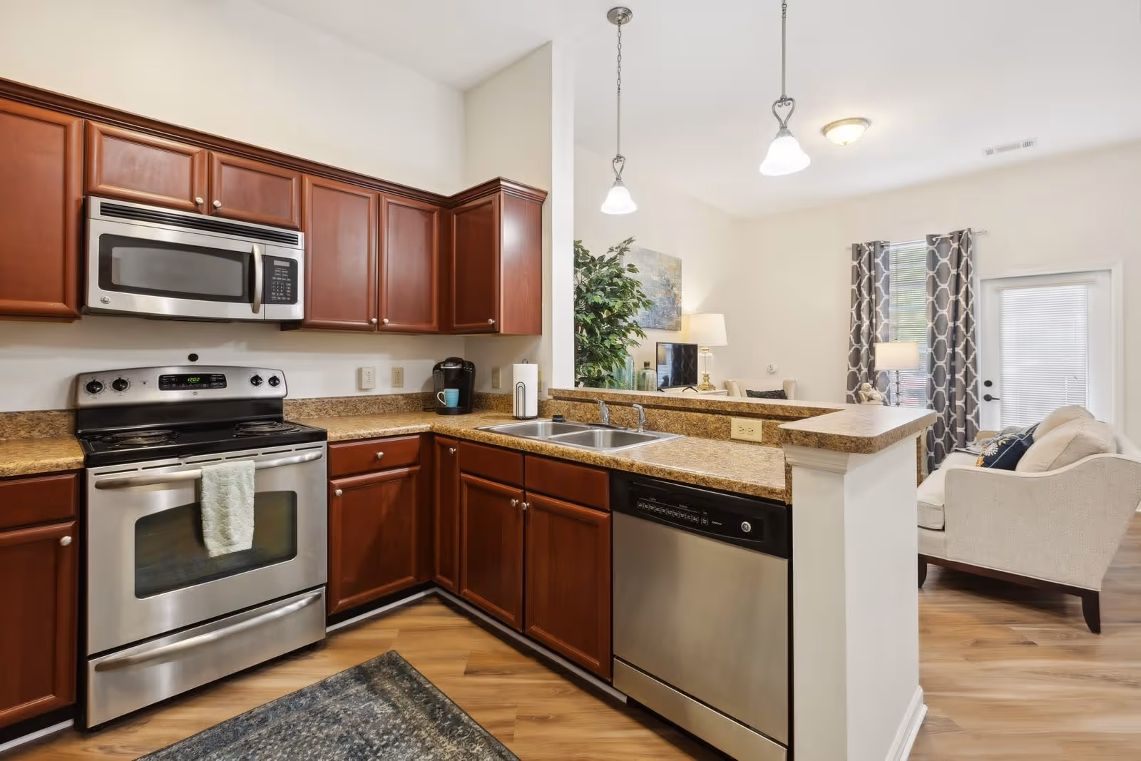 Modern kitchen with stainless steel appliances including a microwave, stove, and dishwasher. The kitchen has wooden cabinets, granite countertops, a double sink, and pendant lights hanging above the counter. In the background, a living room area is visible with a sofa, a lamp, a plant, and a window with patterned curtains.
