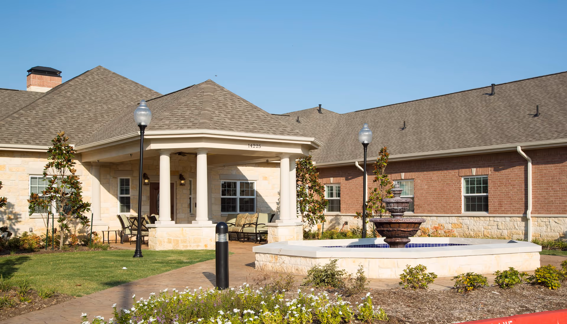 Exterior view of a senior living facility building with a covered porch supported by white columns, outdoor seating, a multi-tiered water fountain in the foreground, landscaped garden beds, and clear blue sky.