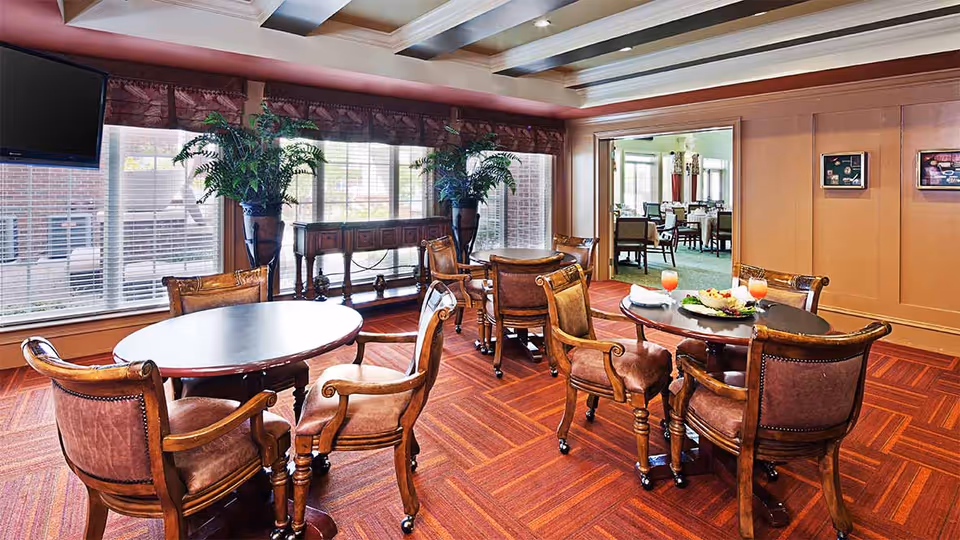 A cozy dining area with round wooden tables and cushioned wooden chairs on a patterned carpet. Large windows with blinds and valances let in natural light. Two large potted plants flank a wooden console table against the window. A flat-screen TV is mounted on the wall to the left. Through an open doorway, another dining room with more tables and chairs is visible.