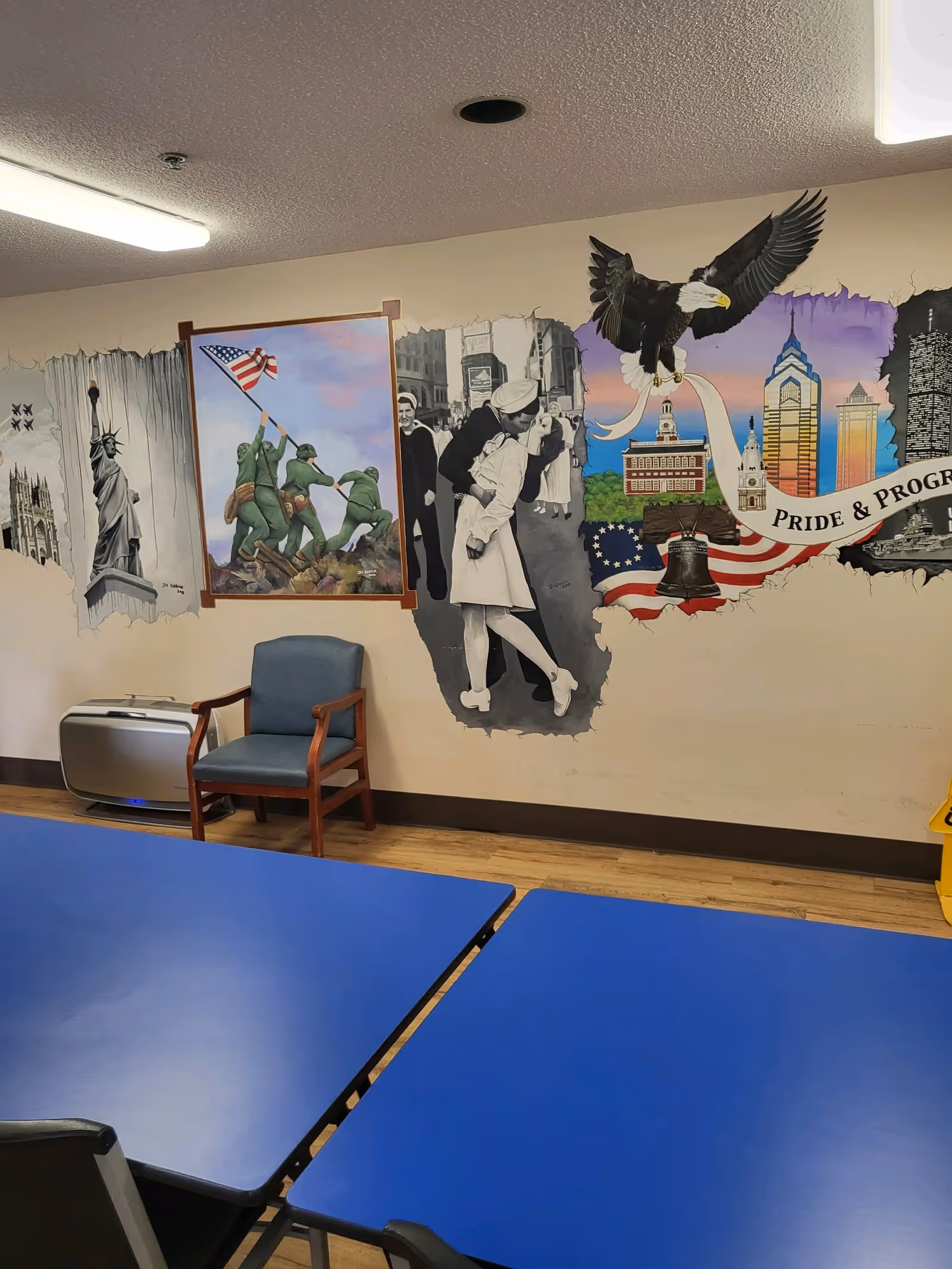 Interior room with blue tables and chairs, a single blue armchair, and a wall mural depicting various patriotic American images including the Statue of Liberty, soldiers raising the flag, a sailor kissing a nurse, an eagle, and notable Philadelphia landmarks with the words 'Pride & Progress'.