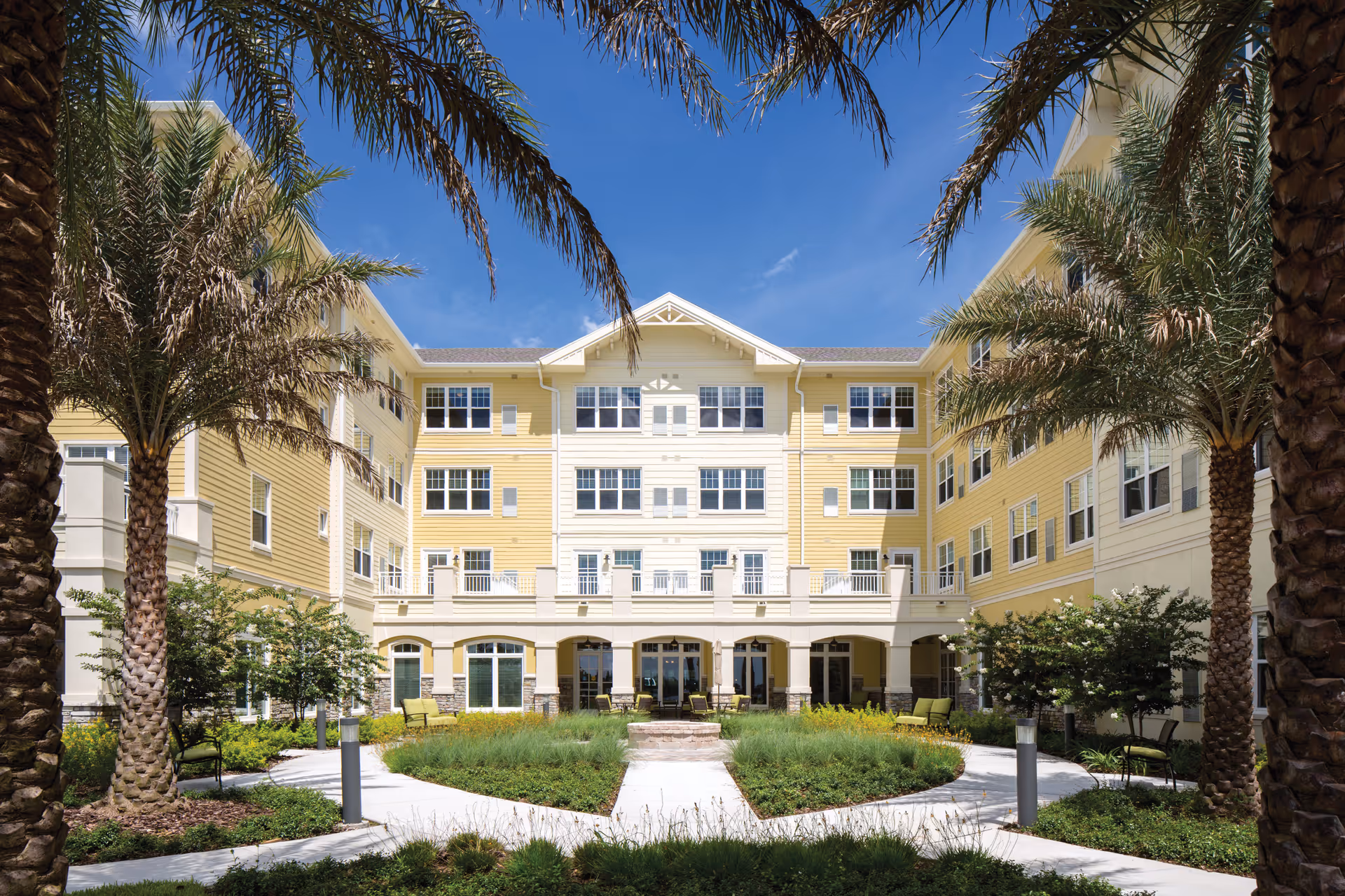 Exterior view of Sonata Windermere senior living facility showing a yellow and white multi-story building with numerous windows. The building surrounds a landscaped courtyard with palm trees, green shrubs, and a paved walkway under a clear blue sky.