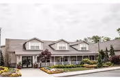 Front exterior of a senior living building with a covered porch, dormer windows, and landscaped flower beds.