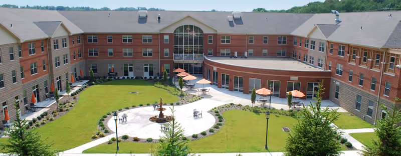 A three-story brick senior living building surrounding a landscaped courtyard with a central fountain, walkways and patio tables with orange umbrellas.