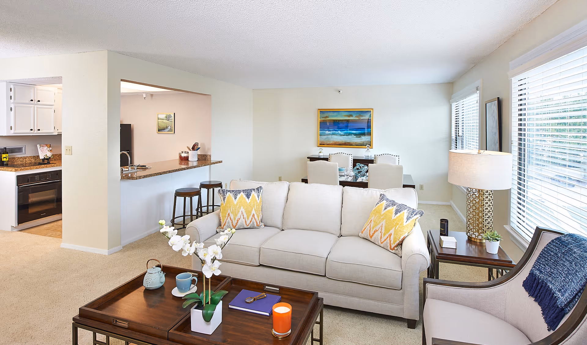 A bright and cozy living room area with a beige sofa adorned with yellow and gray patterned pillows, a wooden coffee table with a white orchid plant, a teapot, a cup, a book, and a candle. To the right is a beige armchair with a blue throw blanket. Behind the sofa is a dining area with a table and chairs, and a colorful painting on the wall. To the left is a kitchen with white cabinets and a granite countertop with bar stools. Large windows with white blinds allow natural light to fill the room.