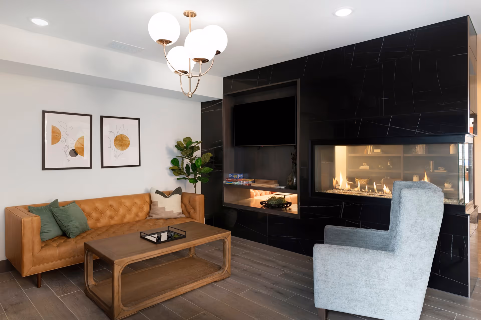 A modern living room with a tan leather tufted sofa adorned with green and beige pillows, a wooden coffee table, a gray upholstered armchair, and a black marble fireplace with a built-in TV above it. Two framed abstract art pieces hang on the white wall, and a green potted plant is placed in the corner. A contemporary chandelier with round white globes hangs from the ceiling.