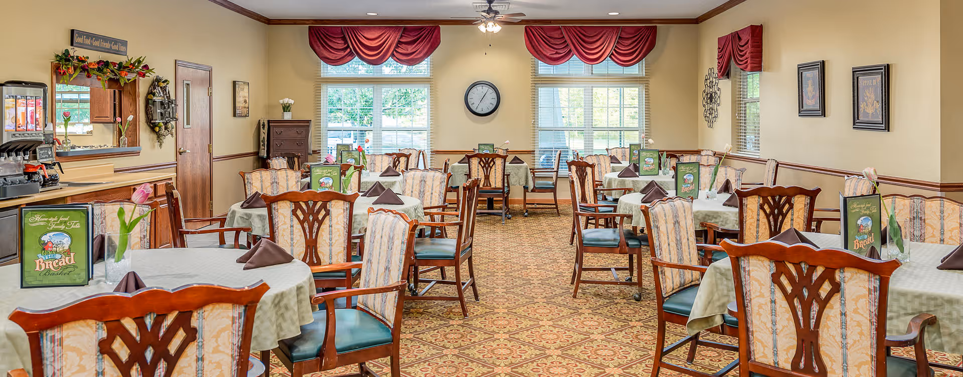 A well-lit dining room with multiple tables covered with light green tablecloths and set with brown napkins and menus. The room features wooden chairs with patterned upholstery, large windows with red valances, a patterned carpet, and a beverage station on the left side. The walls are painted light yellow with wooden trim, and there is a clock centered between the windows.