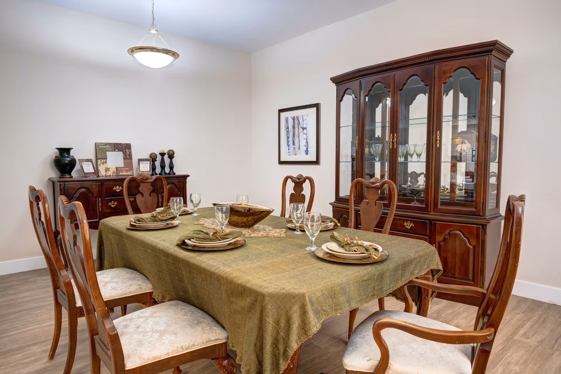 A formal dining room with a wooden dining table covered with a green tablecloth, set with plates, napkins, and glassware for six people. Surrounding the table are six wooden chairs with cushioned seats. Against the wall is a wooden china cabinet with glass doors displaying glassware, and a wooden sideboard with decorative items including a black vase and framed pictures. A hanging light fixture illuminates the room.