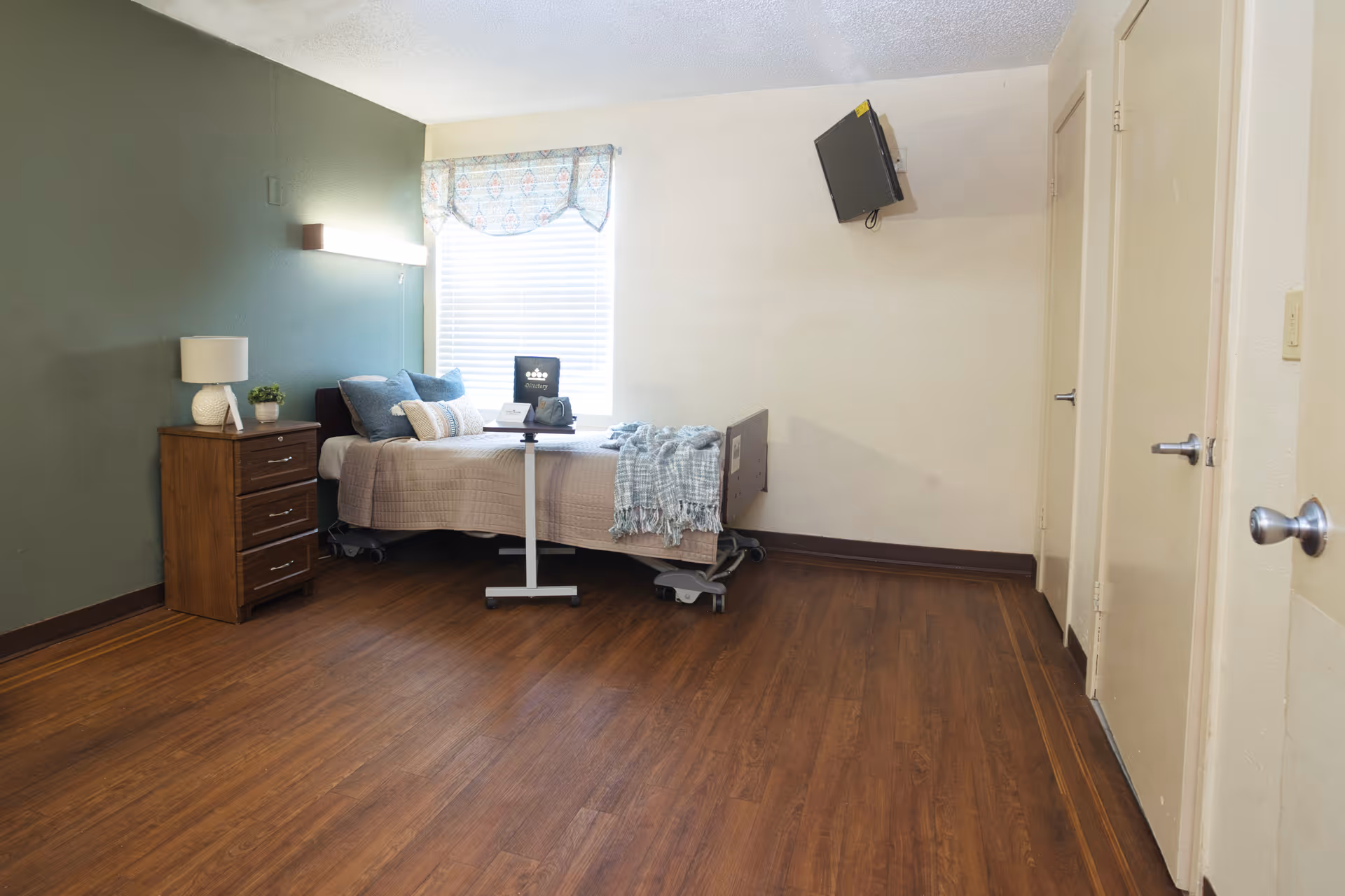 A tidy nursing facility bedroom with a single bed, bedside table and lamp, wall-mounted TV, and wood-look flooring.