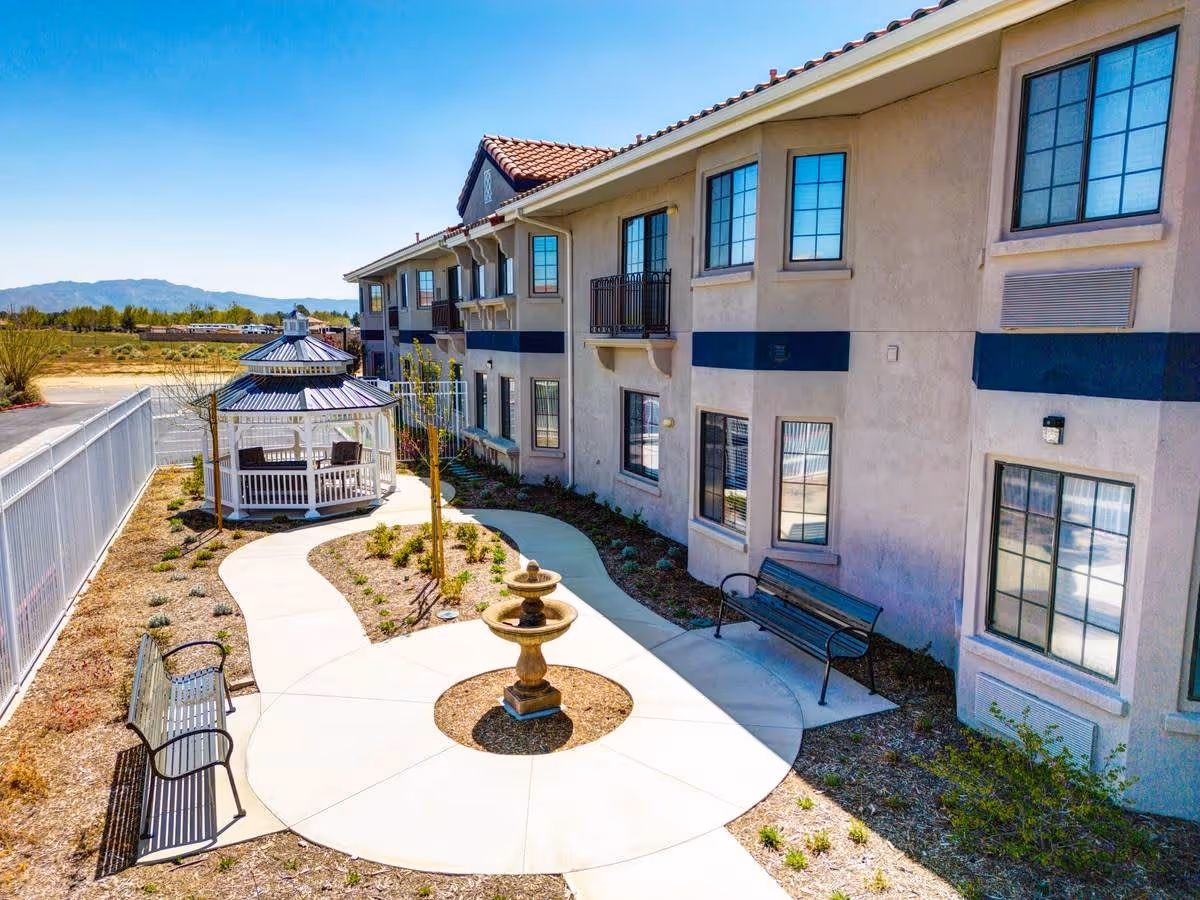 Outdoor courtyard area of a senior living facility with a winding concrete pathway, a central water fountain, benches along the path, and a white gazebo with seating. The building has multiple windows and a beige exterior with a red-tiled roof. Mountains and clear blue sky are visible in the background.