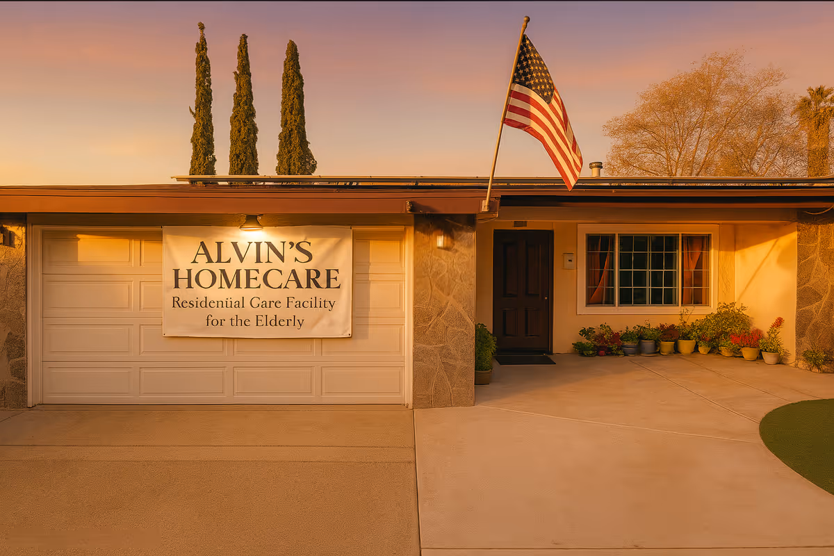 Exterior view of Alvin's Homecare, a residential care facility for the elderly, showing a garage door with a large sign, an American flag mounted on the roof, a front door, a window with curtains, and several potted plants along the walkway.