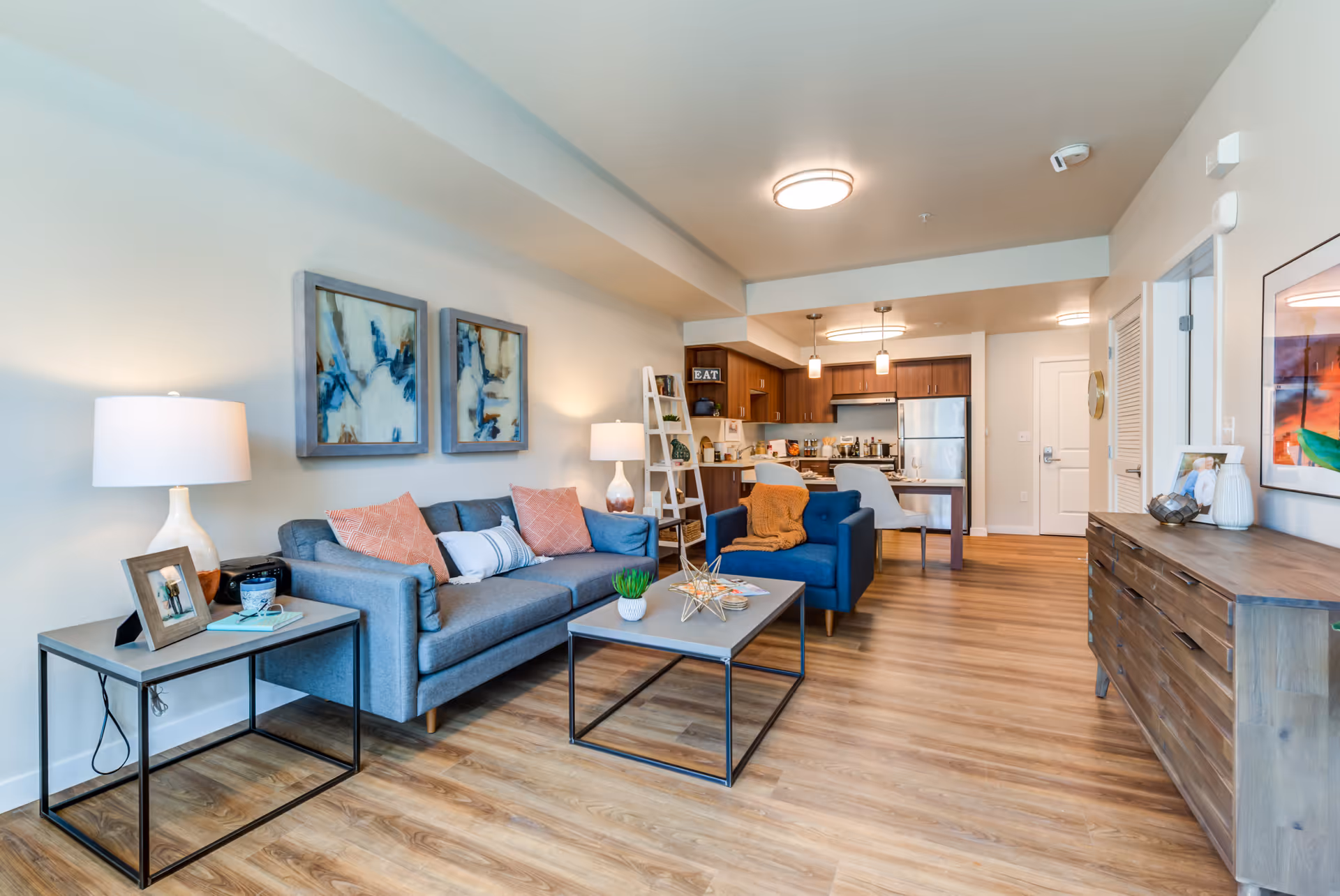 A modern open-concept living room and kitchen area in a senior living facility. The living room features a gray sofa with pink and white pillows, a blue armchair with an orange throw blanket, a coffee table with decorative items, and a side table with a lamp and framed photo. The kitchen has wooden cabinets, stainless steel appliances, and a dining table with two chairs. The space has wood flooring and light-colored walls with abstract artwork.