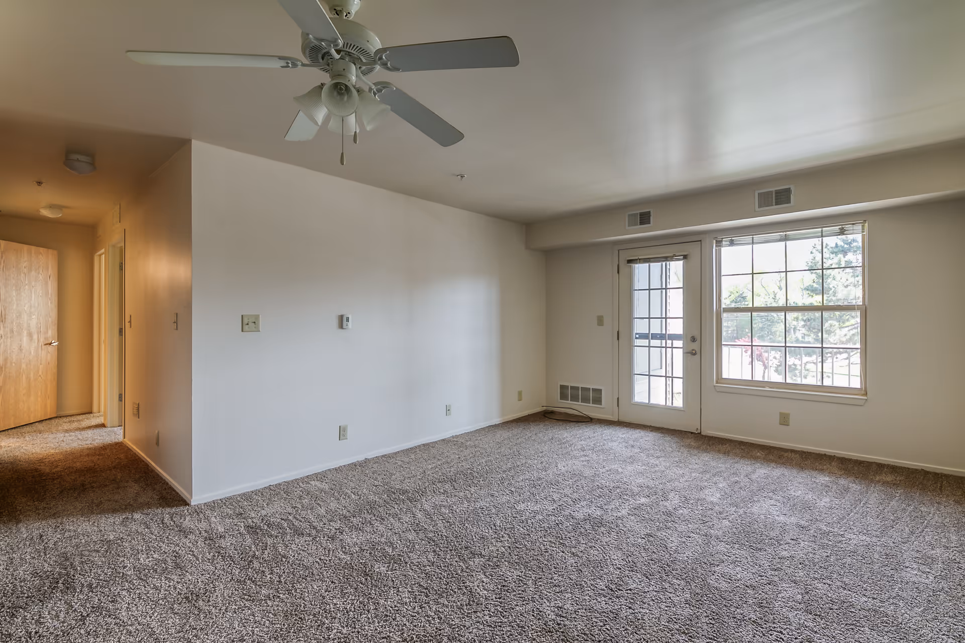 Empty living room with beige carpet, white walls, a ceiling fan, a door with glass panes, and a large window letting in natural light.