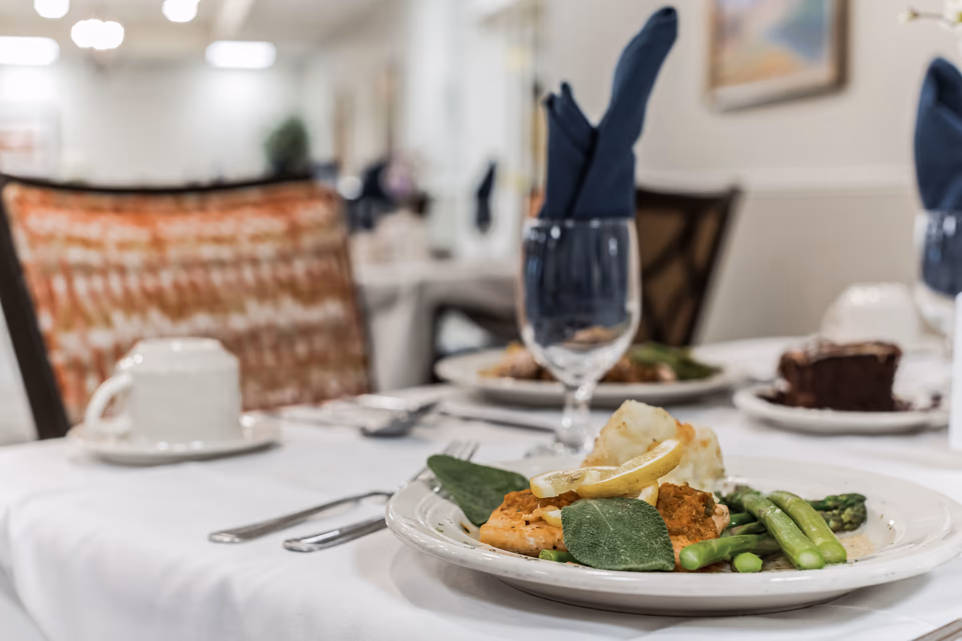 Close-up of a plated meal on a white tablecloth in a dining room setting, featuring asparagus, a lemon garnish, and a main dish with a leafy herb. In the background, there are chairs with patterned cushions, a glass with a folded dark blue napkin, a cup and saucer, and a dessert plate with a slice of chocolate cake.