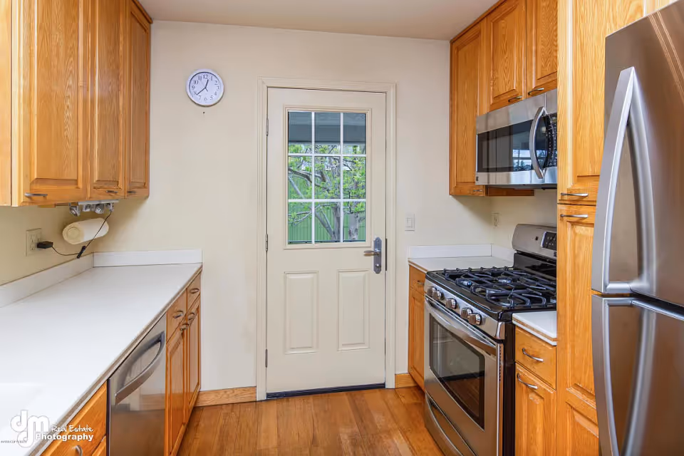 Narrow kitchen with oak cabinets, white countertops, stainless steel appliances and a door with a window to the outside.
