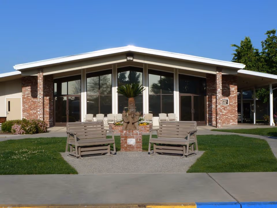 Exterior view of a single-story building with large windows and a peaked roof under a clear blue sky. In front of the building, there are two wooden benches facing each other with a brick planter containing a small palm tree and a sculpture of three elephants. The building has brick and beige walls, and there is a sign on the right side that reads 'DINING ROOM'.