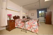 A senior living facility bedroom with two single beds covered in floral bedspreads. Each bed has a bedside table with a lamp and a vase of pink flowers. There is a window with a view of greenery outside, a wooden dresser, and a beige carpeted floor. Privacy curtains are partially drawn around the beds.