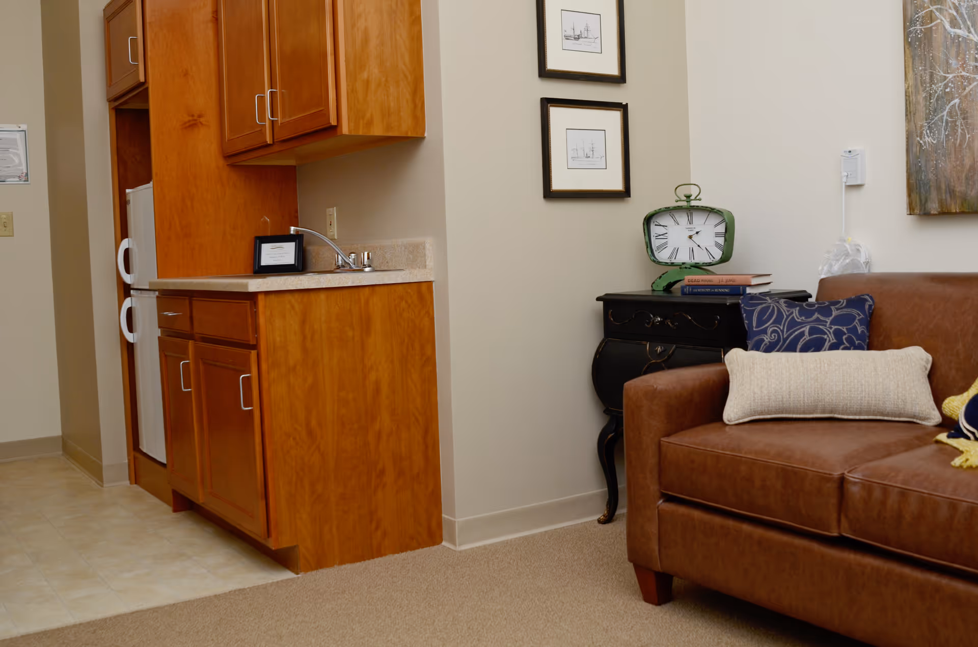 Interior view of a senior living facility room showing a small kitchenette with wooden cabinets and a countertop with a sink. Next to the kitchenette is a brown leather sofa with decorative pillows. A small black side table holds a green clock and some books. Two framed pictures hang on the wall above the side table.