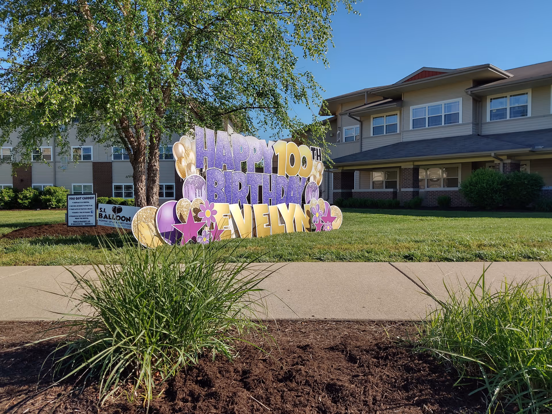 Front lawn of a senior living building with a large colorful 'Happy 100th Birthday Evelyn' yard sign on the grass.