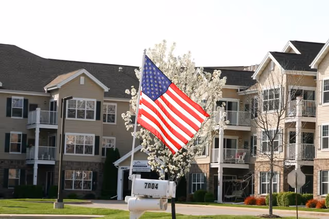 Exterior view of a senior living facility named Parkside Village with beige multi-story buildings, balconies, and a blooming tree with white flowers. An American flag is prominently displayed on a pole near a white mailbox numbered 7004.
