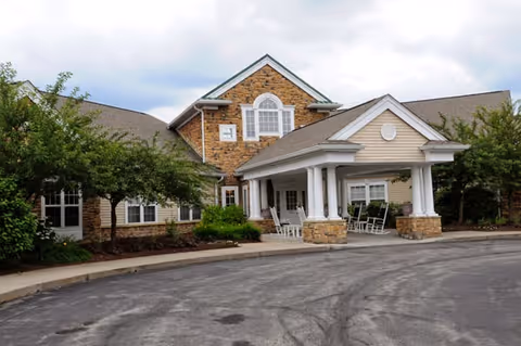 Exterior view of a senior living facility building with a covered entrance supported by white columns. The building features a combination of stone and beige siding, with several windows and a peaked roof. There are trees and shrubs around the entrance and a paved driveway in front.
