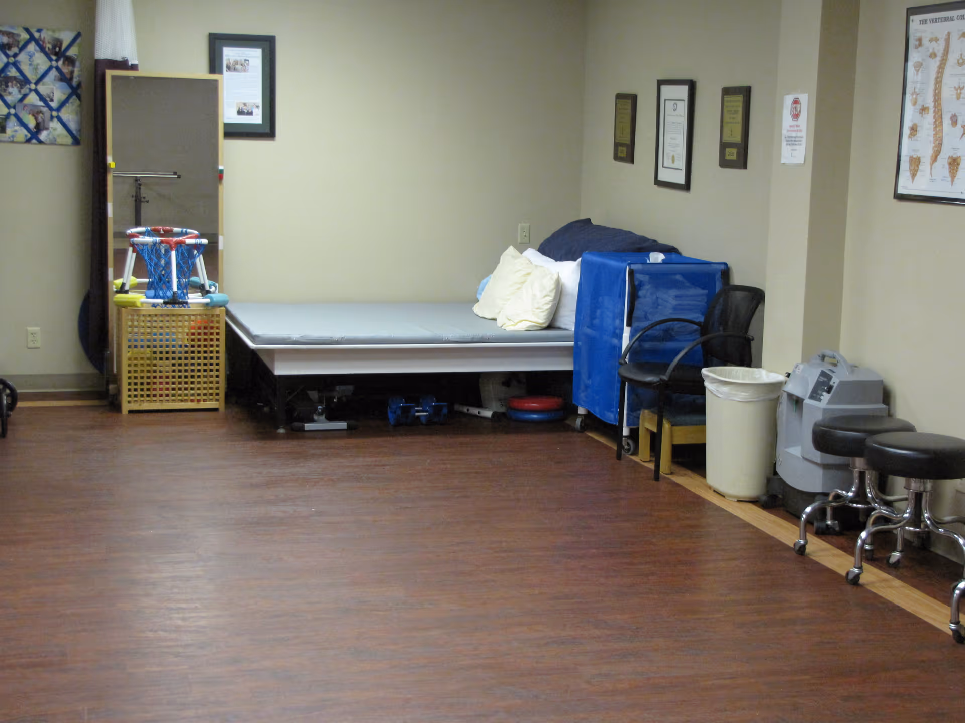 A clinical therapy room with an adjustable treatment bed, chairs, stools, and medical equipment against beige walls and a wood floor.