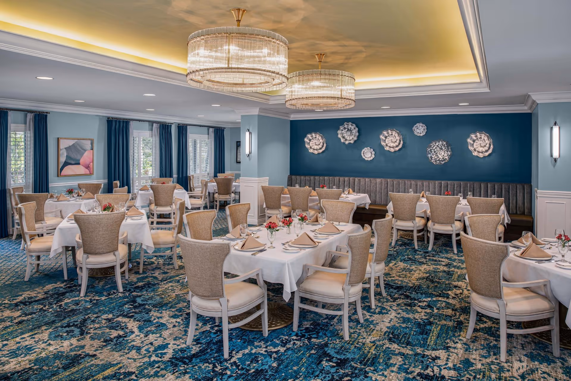 A spacious dining room with multiple tables covered in white tablecloths, each set with beige napkins, glassware, and small floral centerpieces. The room features beige upholstered chairs, blue patterned carpet, blue walls with decorative wall art, large windows with blue curtains, and two elegant circular chandeliers hanging from a recessed ceiling with soft lighting.