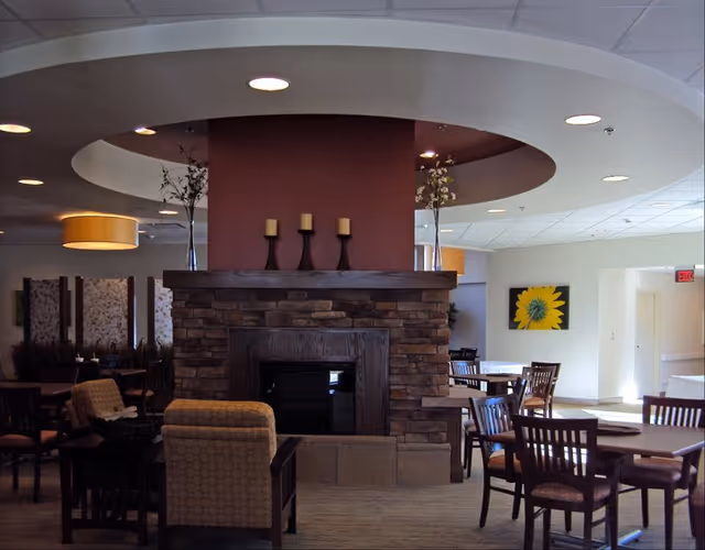 A cozy common area in a senior living facility featuring a stone fireplace with three candles on the mantel. The room has several wooden tables and chairs arranged for dining or socializing. There are comfortable armchairs near the fireplace, decorative plants, and a large sunflower painting on the wall. The ceiling has recessed lighting and a circular architectural feature above the fireplace.