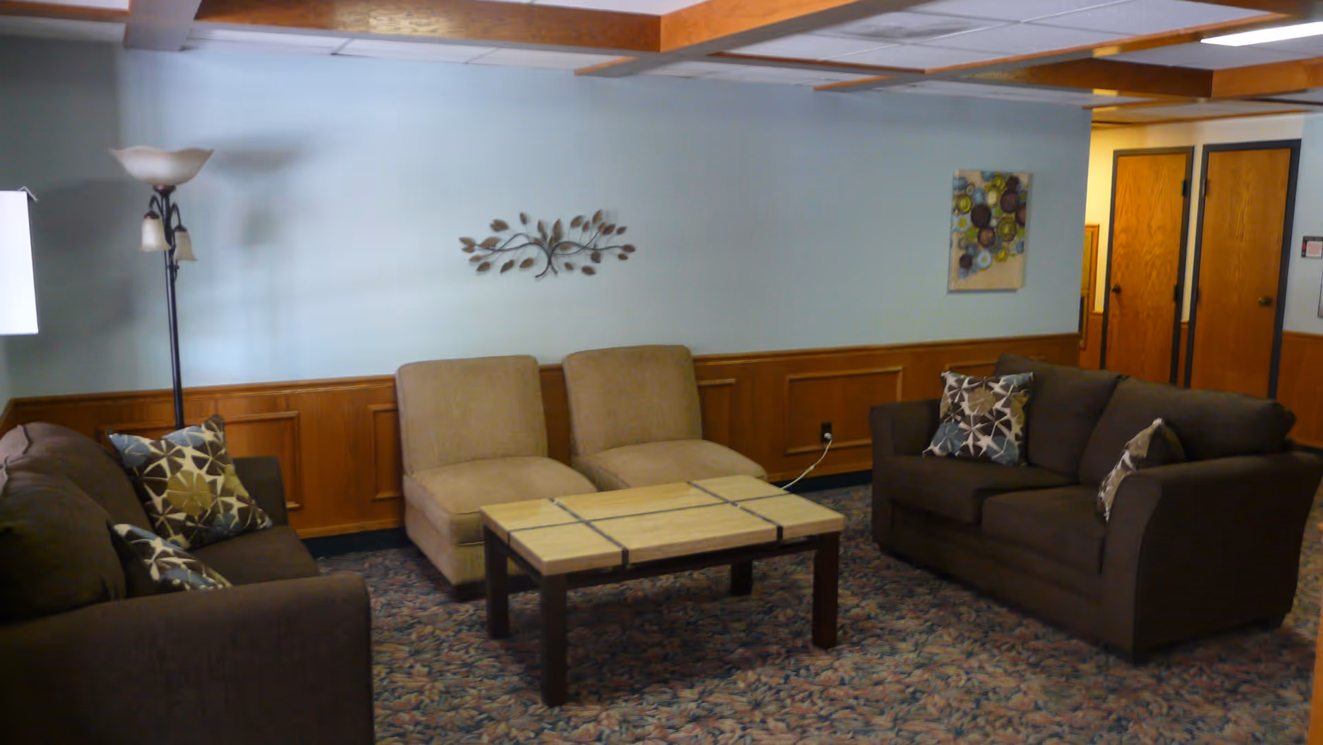 A cozy living room with two brown sofas adorned with patterned pillows, two beige chairs, a rectangular coffee table with a tiled top, a floor lamp, and wall decorations including a metal leaf sculpture and a colorful abstract painting. The room has wood paneling on the lower half of the walls and a patterned carpet.