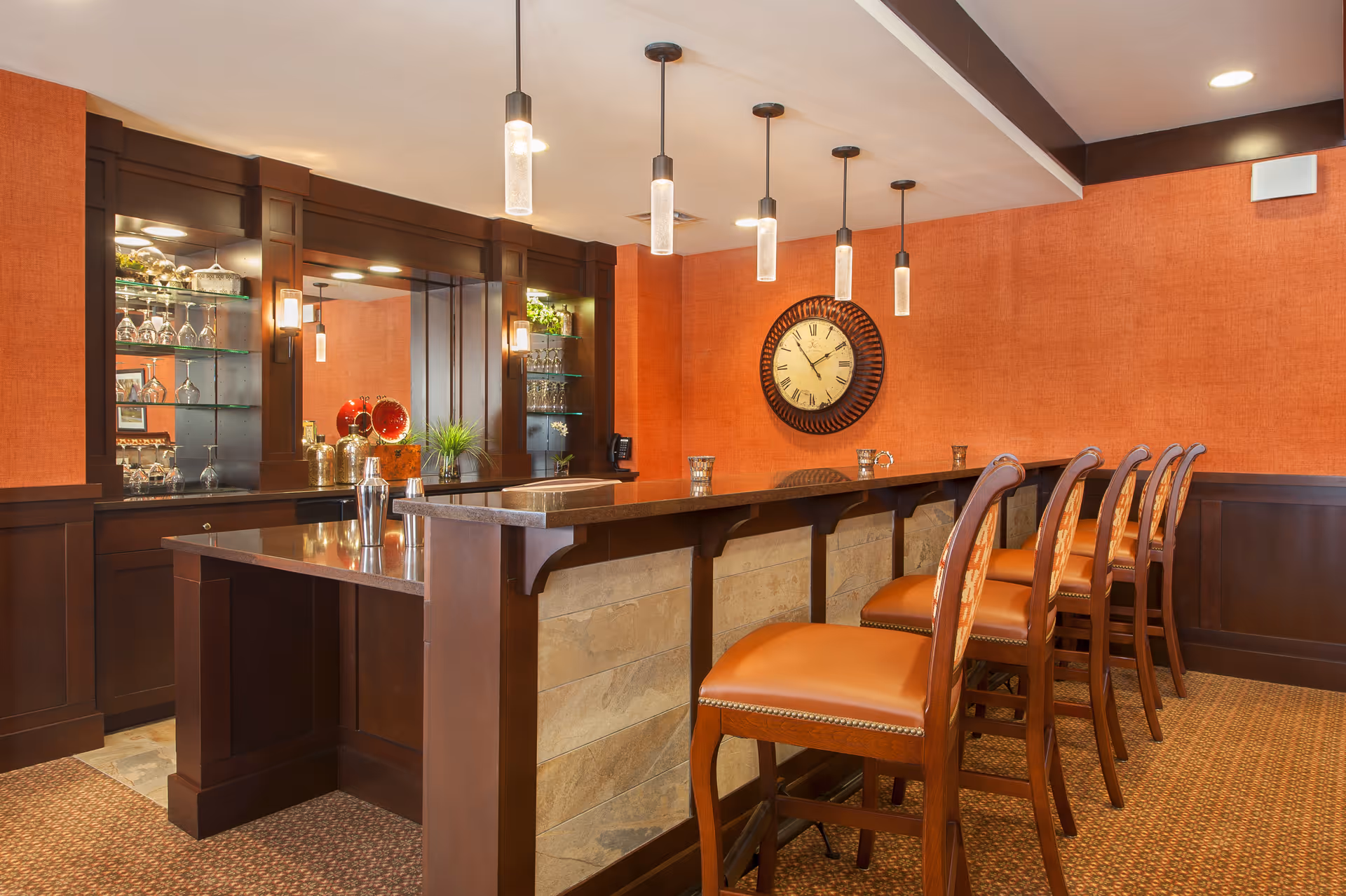 Interior view of a senior living facility's bar area with a long counter, five high wooden chairs with orange cushions, pendant lights hanging from the ceiling, a large round clock on an orange textured wall, and shelves with glassware and decorative items.