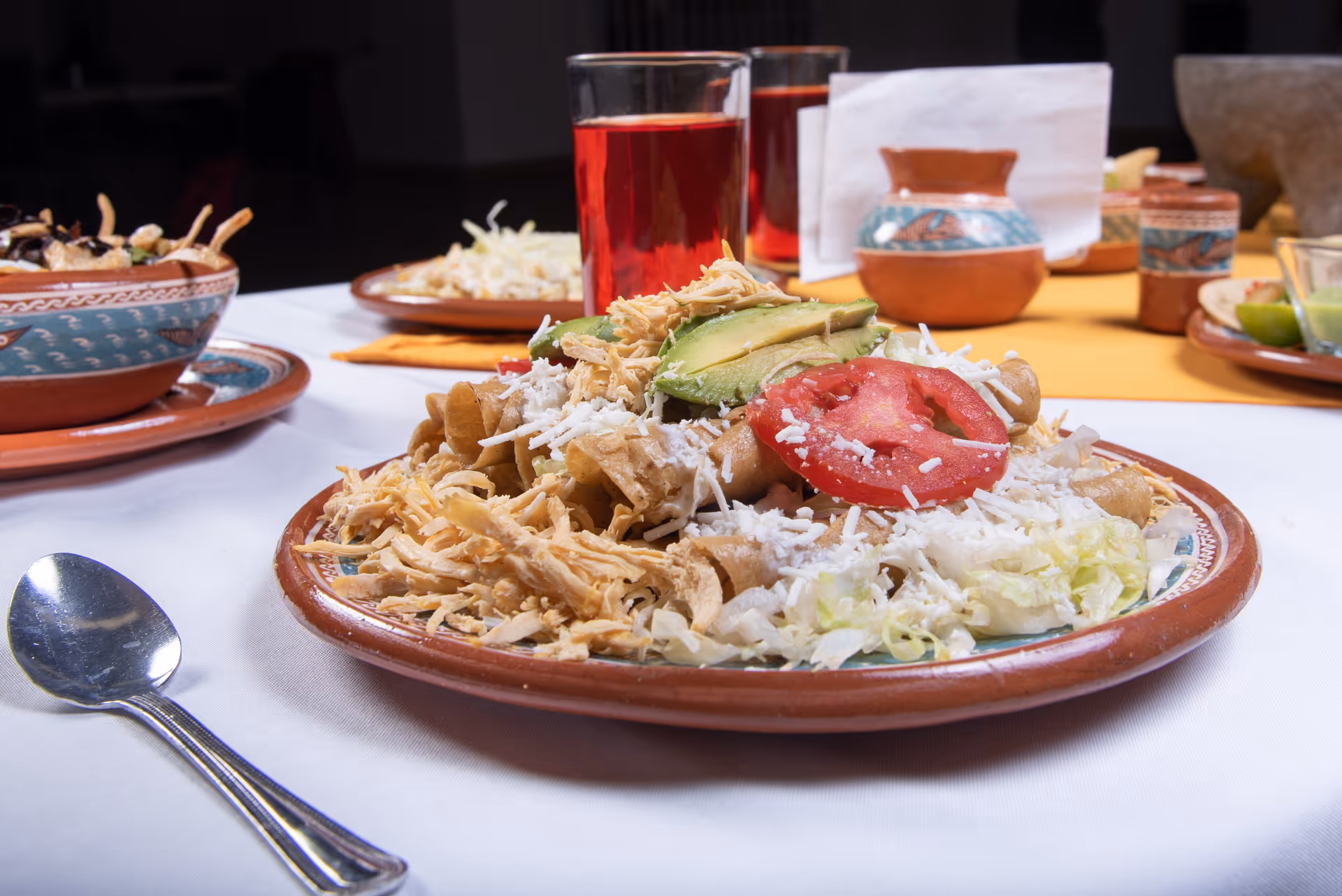 A close-up view of a plate of food on a white tablecloth, featuring shredded chicken, sliced avocado, tomato, lettuce, and cheese on a traditional ceramic plate. In the background, there are two glasses filled with a red beverage and additional plates and ceramic containers on the table.