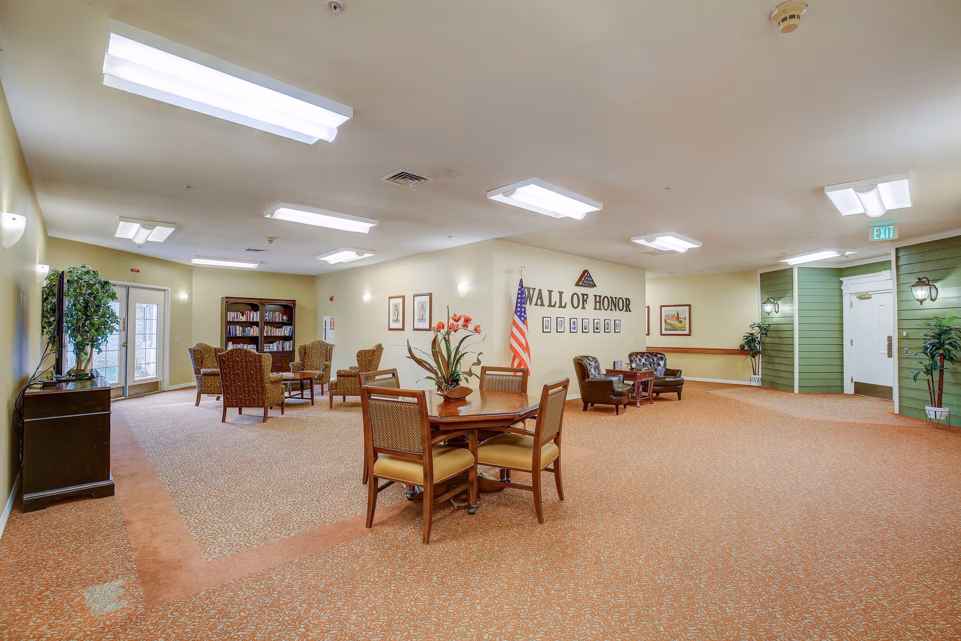 A spacious senior living facility common area with carpeted floors and beige walls. There are several seating arrangements including a round wooden table with four chairs in the foreground, a group of four upholstered armchairs around a small table near a bookshelf filled with books, and two leather chairs with a small table near a wall labeled 'Wall of Honor' with framed photos and an American flag. The room is well-lit with ceiling lights and has exit doors on the left and right sides.